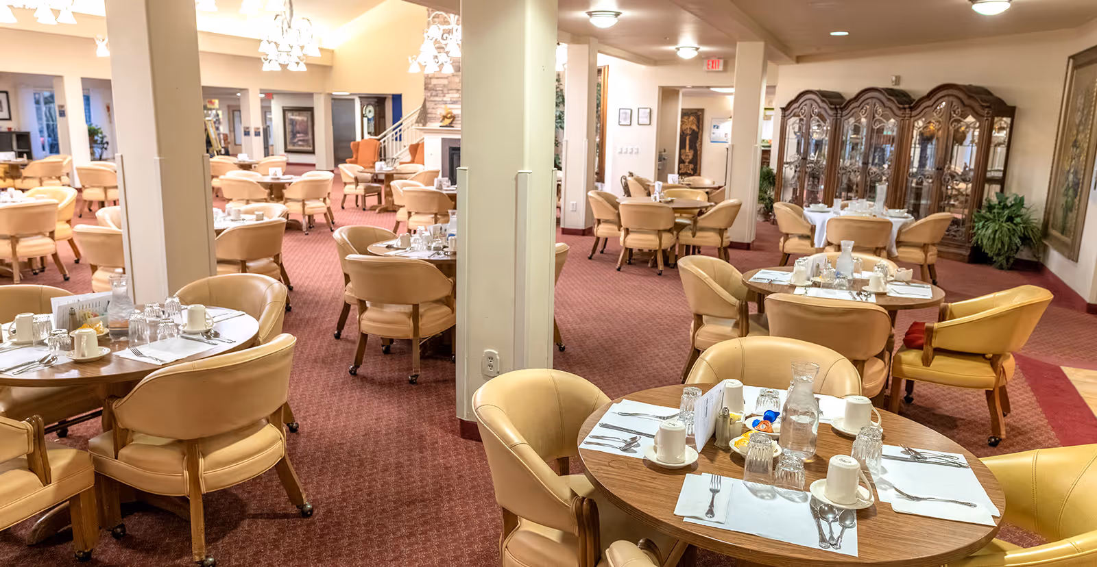 Spacious dining room with round tables set for meals, beige chairs, chandeliers, and a china cabinet in the background.