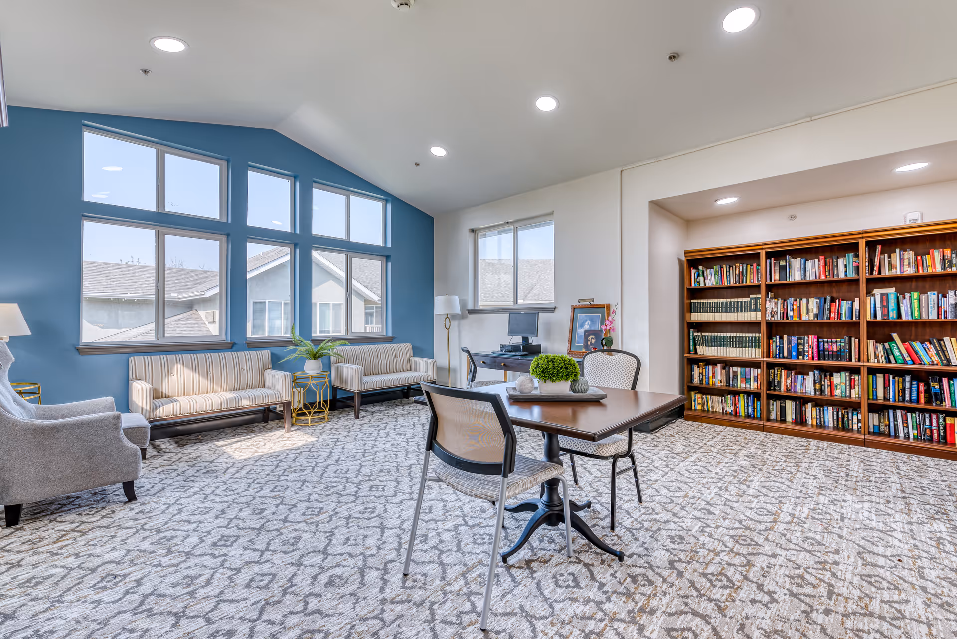 A bright and spacious room with large windows allowing natural light to fill the space. The room features a patterned carpet, two striped sofas against a blue accent wall, a gray armchair, a wooden table with two chairs, and a large bookshelf filled with books. There is also a desk with a computer and a framed photo near the window.