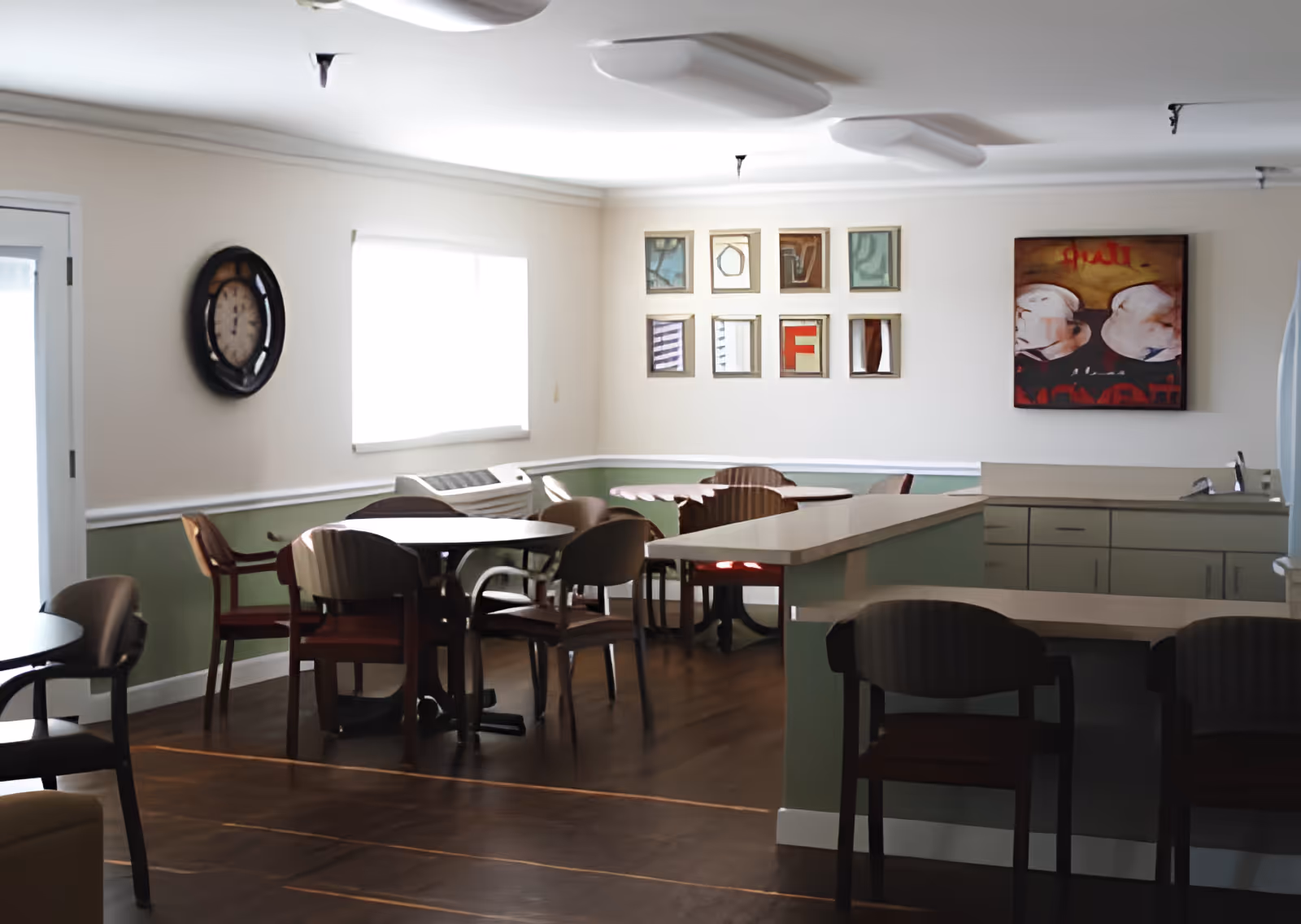 Communal dining area with tables, chairs, and a serving counter/kitchenette decorated with wall art and a clock.