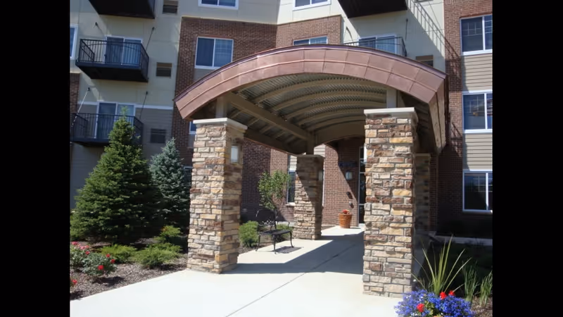 Entrance to a multi-story residential building with a covered walkway supported by stone pillars. There are benches along the walkway, landscaped greenery including bushes, flowers, and small evergreen trees around the entrance area.
