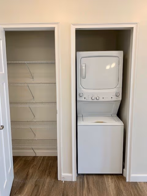 Stacked washer and dryer in a closet next to an open pantry with wire shelving on wood-look flooring.