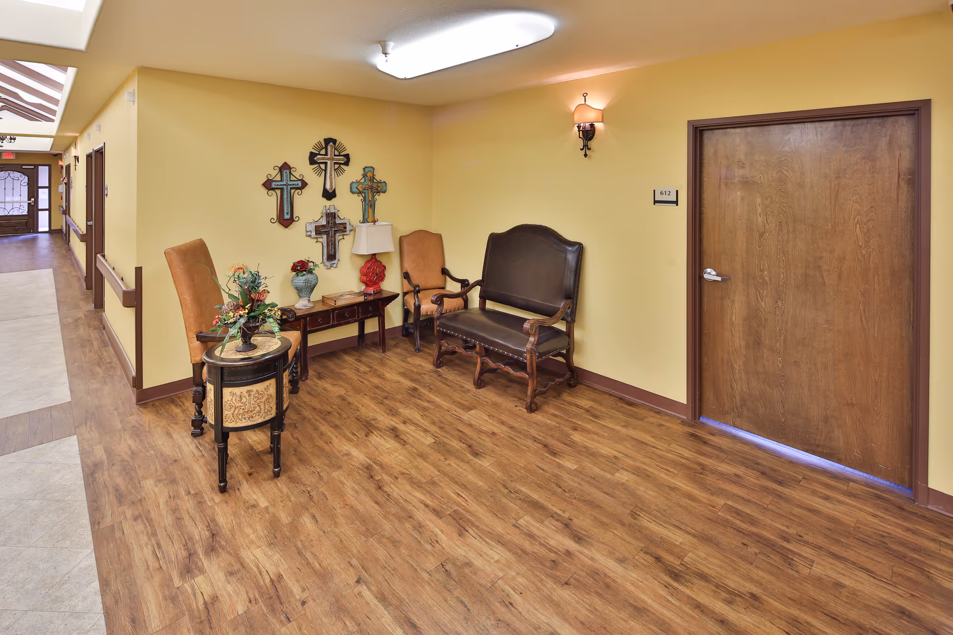 Hallway seating area in a senior living facility with chairs, small tables and decorative wall crosses next to a closed wooden door numbered 612.
