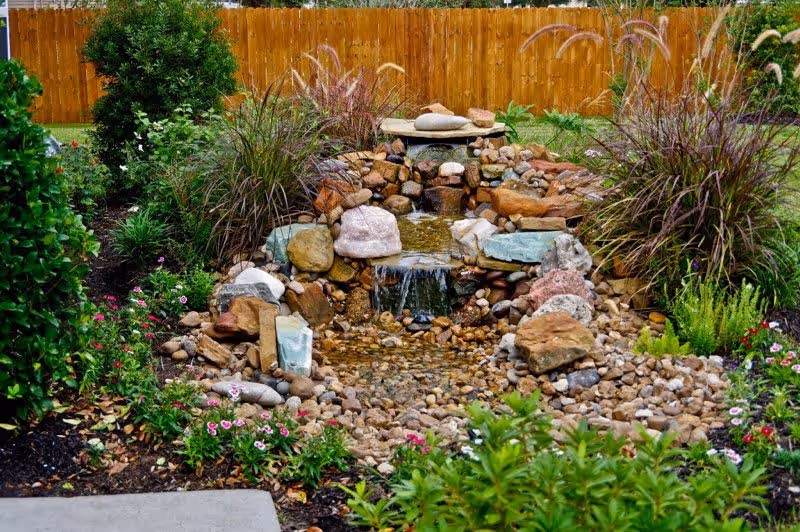 A landscaped garden area featuring a small cascading rock waterfall surrounded by various plants, flowers, and shrubs, with a wooden fence in the background.