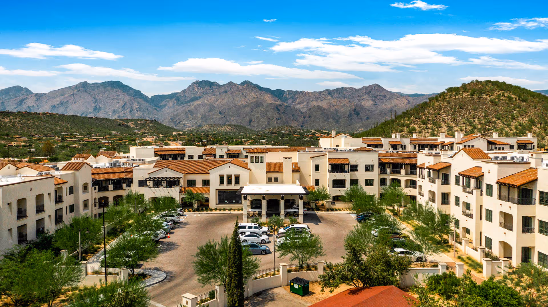Aerial view of The Hacienda at the Canyon senior living facility with multiple beige buildings featuring red-tiled roofs, surrounded by desert landscaping and parking areas. In the background, there are rugged mountains under a partly cloudy blue sky.