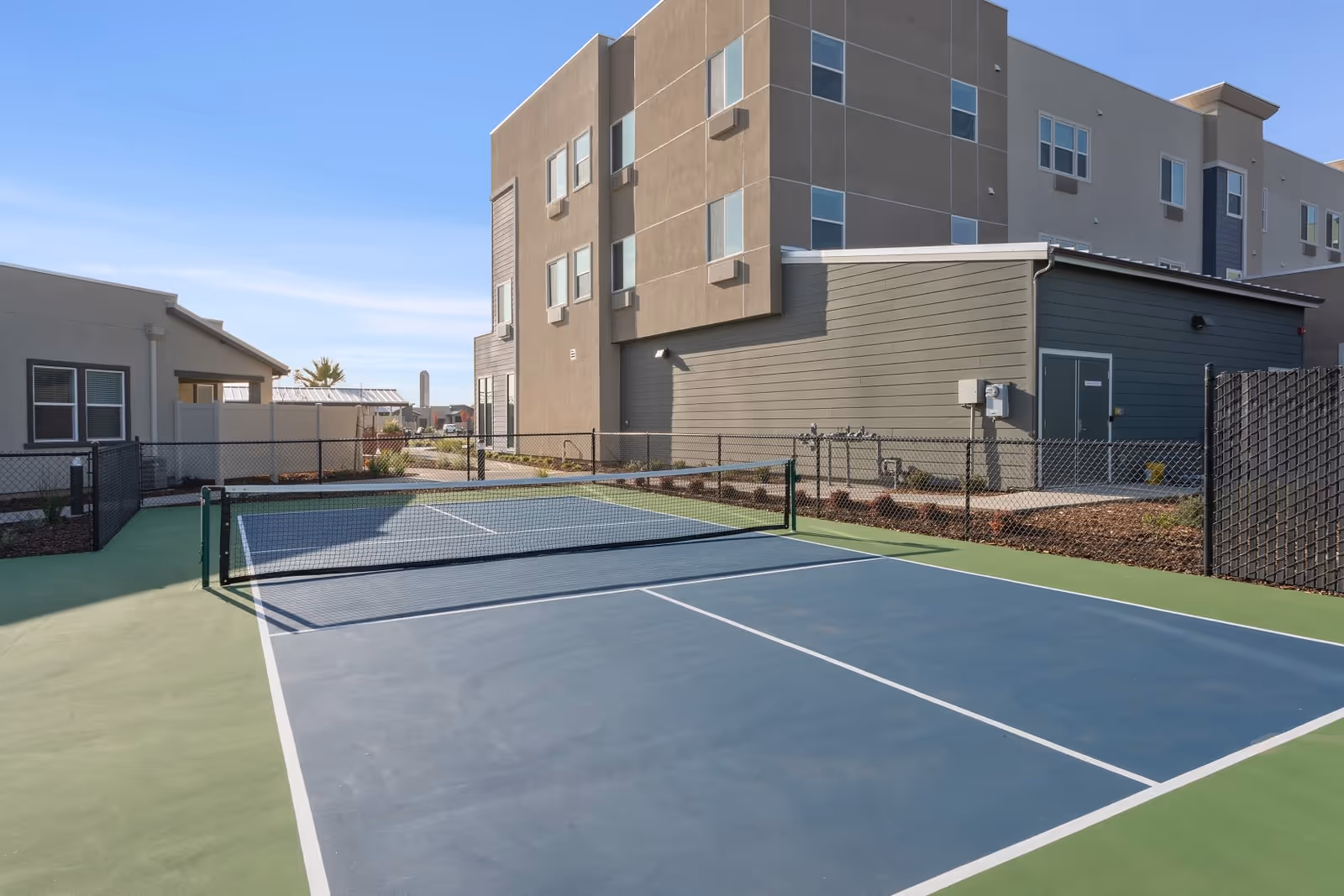A fenced outdoor tennis/pickleball court beside a multi-story residential building under a clear sky.
