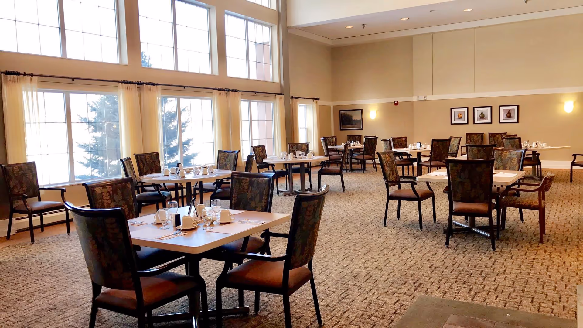 A spacious dining room with multiple tables and chairs arranged neatly. Each table is set with cups, glasses, and utensils. Large windows with sheer curtains allow natural light to fill the room. The walls are decorated with framed artwork and soft wall lights.