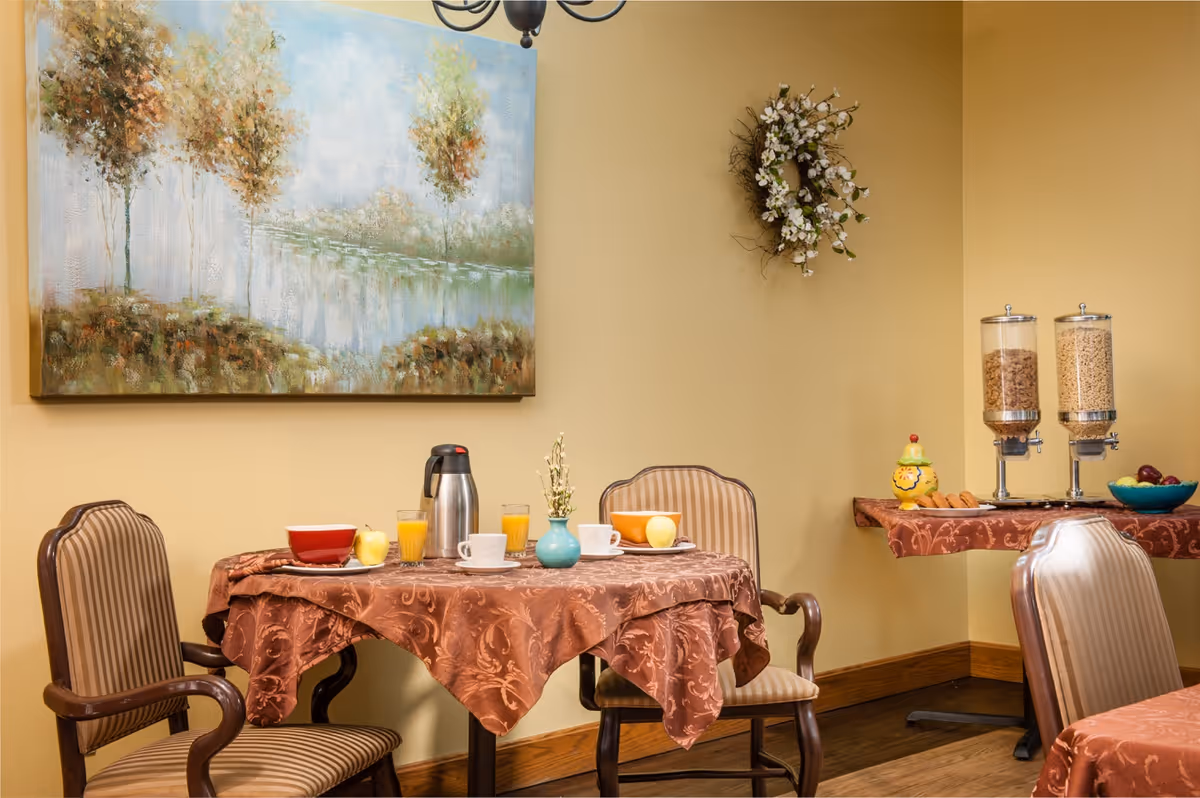 A cozy dining area with a round table covered by a brown patterned tablecloth. The table is set with two bowls, two glasses of orange juice, a coffee pot, two cups, and two apples. Two striped upholstered chairs are positioned around the table. On the wall above the table is a large painting of trees by a lake and a decorative floral wreath. To the right, a side table holds two cereal dispensers, a small decorative container, and a bowl of fruit.