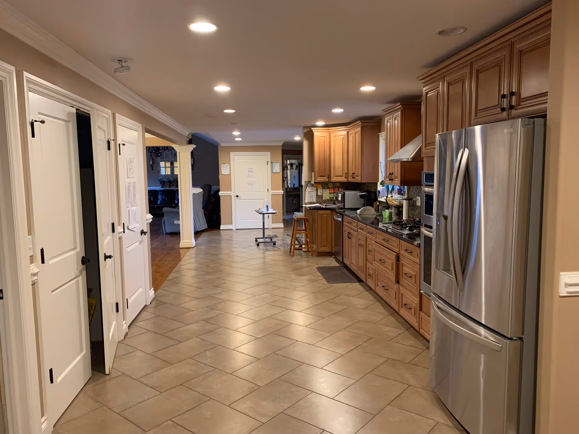 A long, spacious kitchen area with tiled floor and wooden cabinets on the right side. The kitchen features a stainless steel refrigerator, built-in oven, stovetop with a range hood, and various kitchen appliances on the countertop. On the left side, there are several white doors and an open doorway leading to a living room area with chairs and a sofa. The ceiling has recessed lighting evenly spaced along the length of the room.