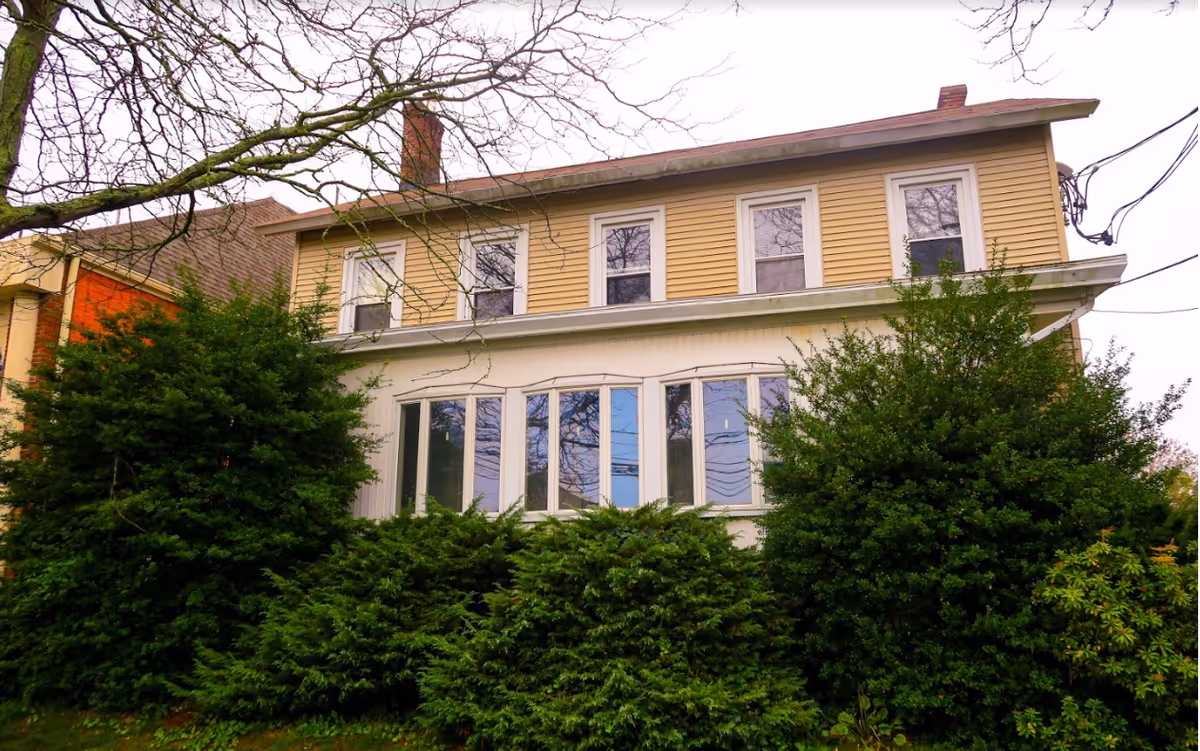 Exterior view of a two-story residential building with beige siding and multiple windows, surrounded by green bushes and trees with bare branches.