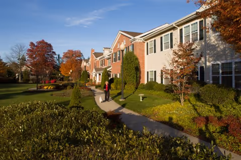Well-maintained senior living building exterior with a landscaped walkway and two people walking under a blue sky.
