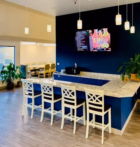Interior view of a modern common area in a senior living facility featuring a blue and white kitchen island with four white bar stools, a mounted TV on a dark blue wall displaying colorful text, pendant lights hanging from the ceiling, and a dining area with tables and chairs visible in the background.