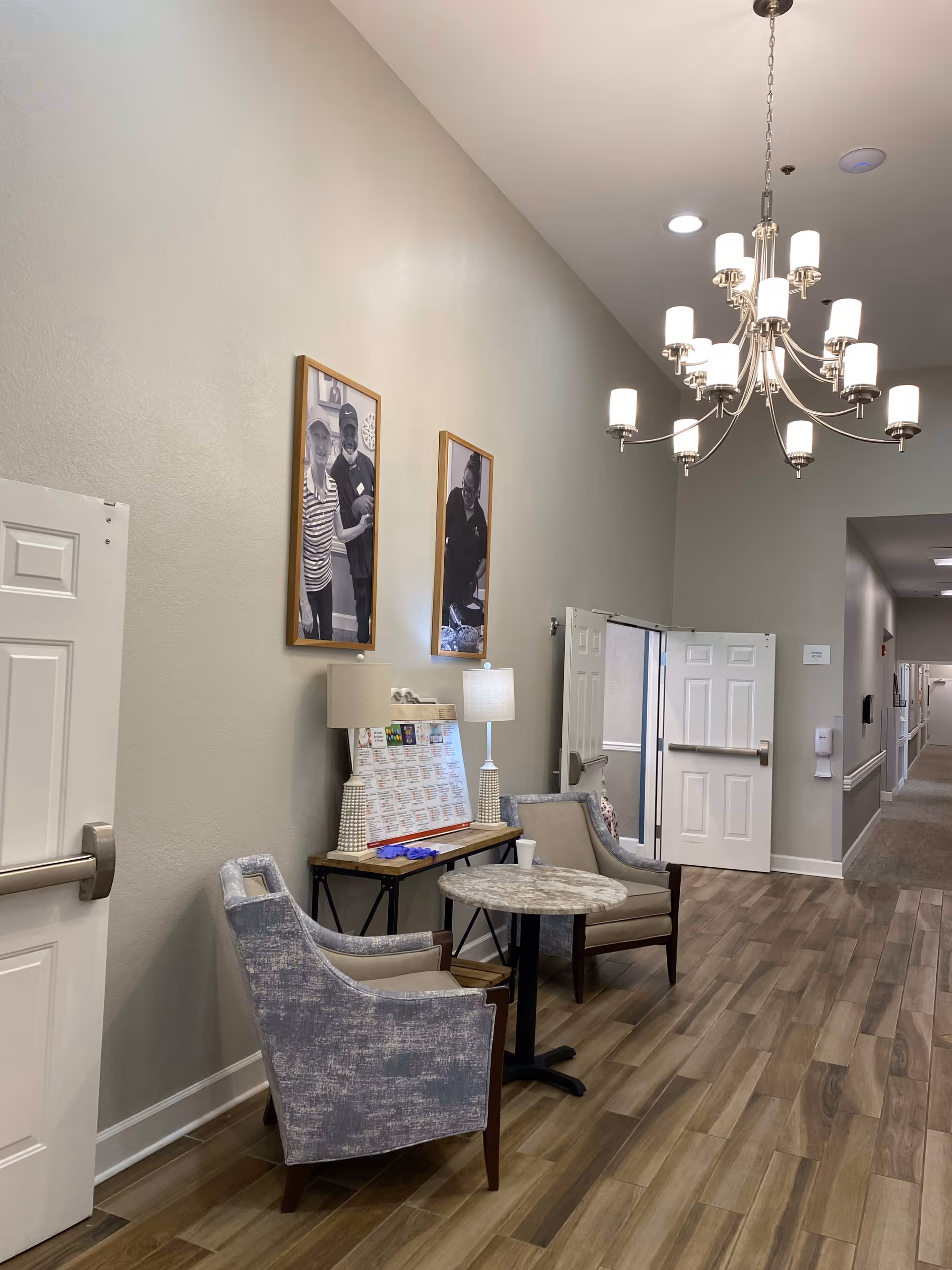 A hallway in a senior living facility with two upholstered chairs and a small round table between them. A console table with two lamps and a calendar is against the wall. Two framed black and white photos hang above the table. The floor is wood-patterned, and a modern chandelier hangs from the ceiling. Doors and a long corridor are visible in the background.