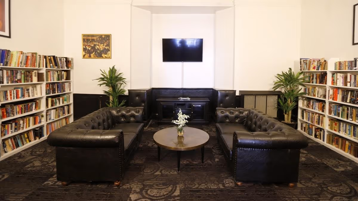 A cozy living room area with two black leather tufted sofas facing each other across a round wooden coffee table with a small flower arrangement. The room has bookshelves filled with books on both sides, two green potted plants, a wall-mounted flat screen TV, and a framed picture on the wall.