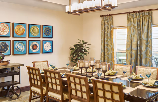 A well-lit dining room with a long wooden table set for eight people. The table is decorated with green napkins, blue water glasses, and a centerpiece of multiple candle holders. The room has patterned curtains on the window, a potted plant in the corner, and a wall display of framed decorative plates.
