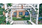 A white wooden garden arbor with lattice sides and a bench underneath, surrounded by plants and greenery. In the background, there is a brick building with windows and a small garden area with a bench and flowers.
