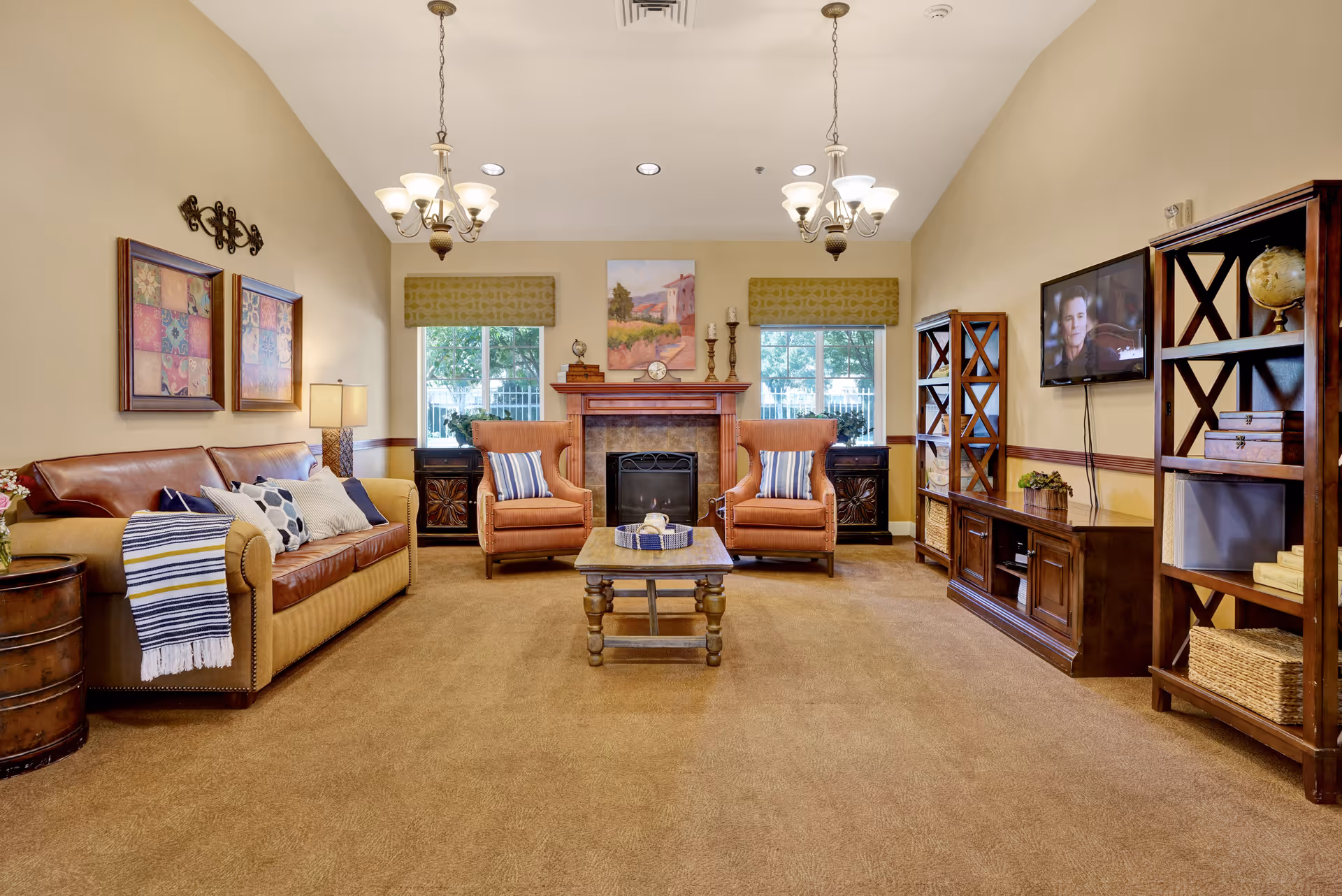 A cozy living room with a beige carpet and walls, featuring a brown leather sofa with multiple cushions and a striped throw blanket on the left. Two orange armchairs with striped pillows are positioned in front of a fireplace with a wooden mantel, which has decorative items and a painting above it. There are two windows with green valances on either side of the fireplace. On the right side, there is a wooden entertainment center with a flat-screen TV mounted on the wall and two matching wooden bookshelves with decorative items. Two chandeliers hang from the ceiling providing warm lighting.