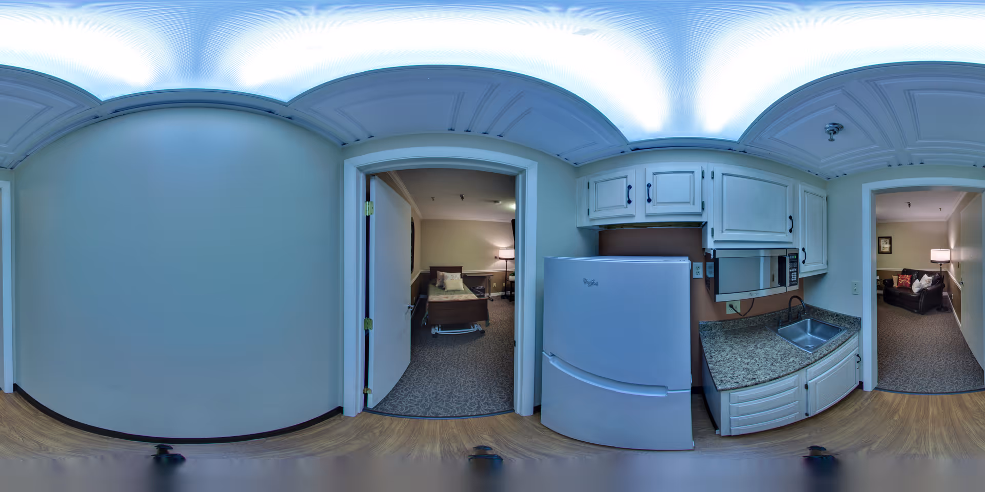 A panoramic view of an interior hallway in a senior living facility. On the left, there is an open door leading to a bedroom with a bed, nightstand, and lamp. In the center, there is a kitchenette area with white cabinets, a refrigerator, a microwave, and a sink with granite countertops. On the right, another open doorway leads to a living room area with a sofa and a lamp. The floor is wood laminate, and the ceiling has decorative paneling with bright lighting.