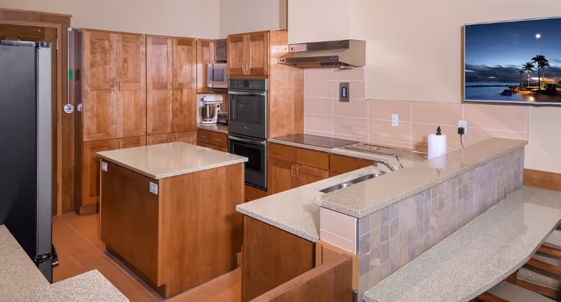 A modern kitchen with wooden cabinets, a central island with a beige countertop, built-in double ovens, a stainless steel refrigerator, a sink, and a mounted flat-screen TV displaying a scenic beach sunset with palm trees.