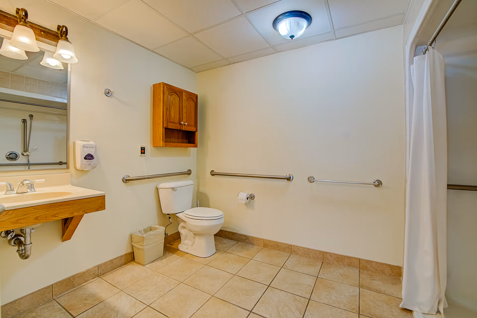 A spacious bathroom with beige tiled floor and white walls. It features a white toilet with grab bars on the walls around it, a wooden cabinet mounted above the toilet, a sink with a wooden vanity and a mirror above it, a soap dispenser on the wall, a trash bin below the sink, and a shower area with a white curtain on the right side.