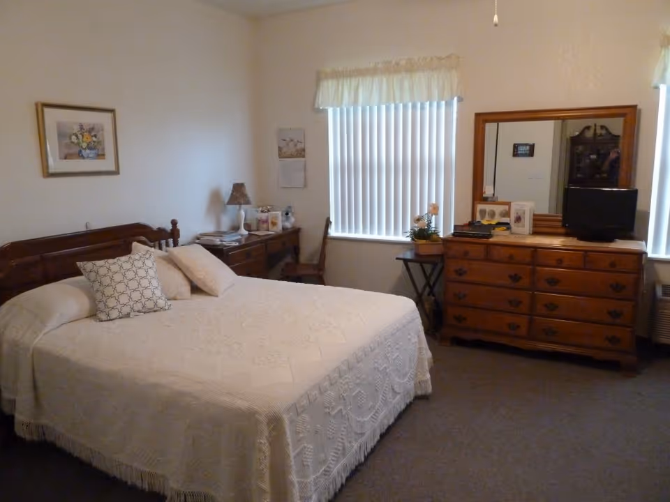 Furnished bedroom with a bed covered in a white bedspread, wooden dresser with mirror, desk, and a window with vertical blinds.