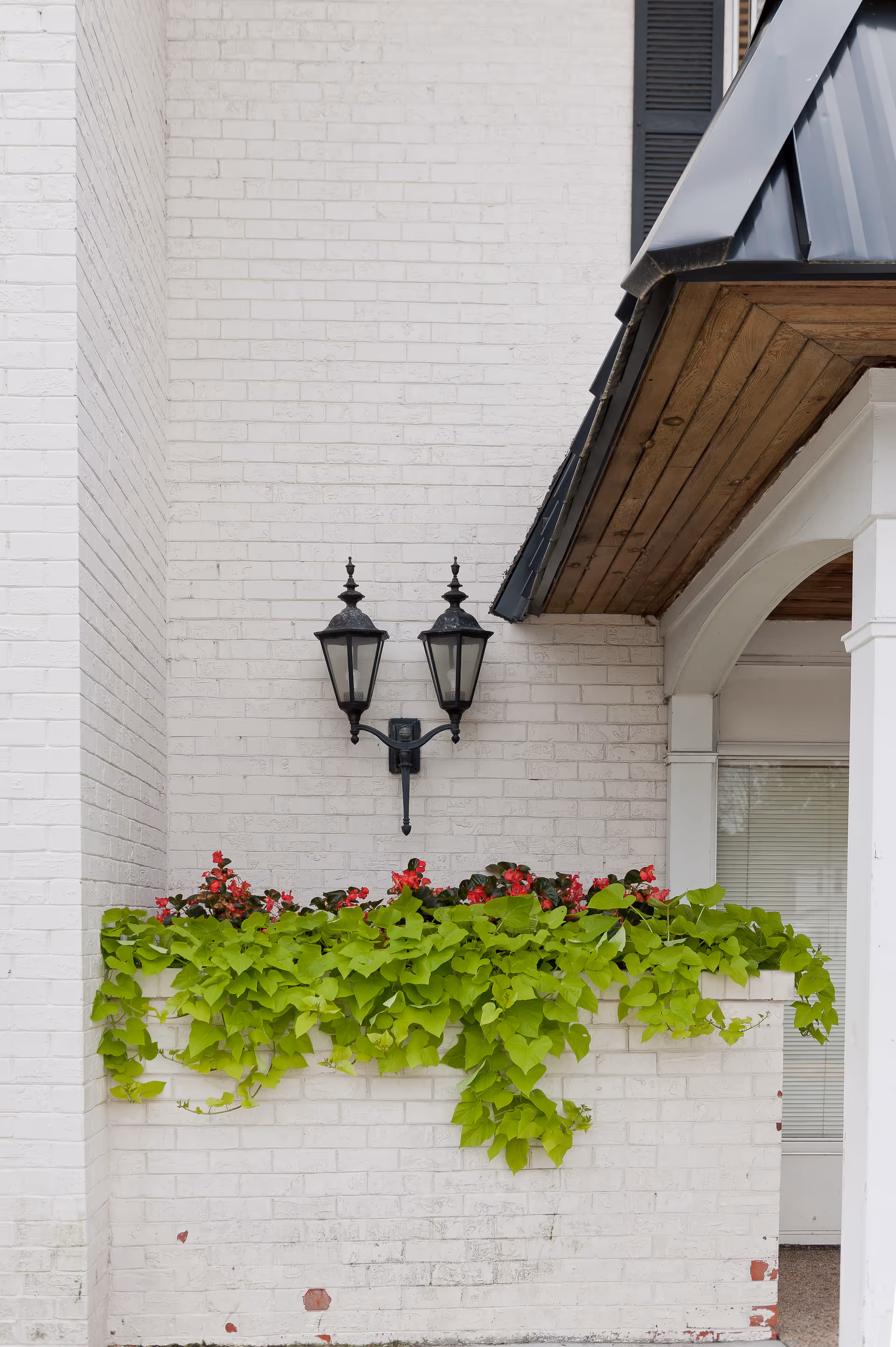 White brick exterior wall with a black double lantern-style light fixture mounted above a planter box filled with green leafy plants and red flowers. Part of a roof with wooden underside and white columns is visible on the right side.