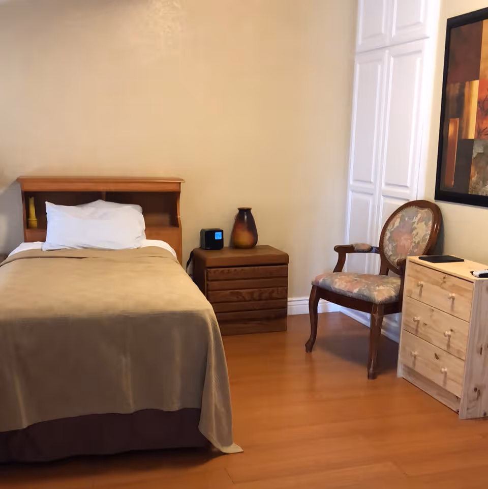 A simple bedroom with a single bed covered with a beige blanket and a white pillow. Next to the bed is a wooden nightstand with a digital clock and a decorative vase. There is a floral upholstered wooden chair and a small wooden dresser with drawers. A framed abstract painting hangs on the wall above the dresser. The room has wooden flooring and light-colored walls.