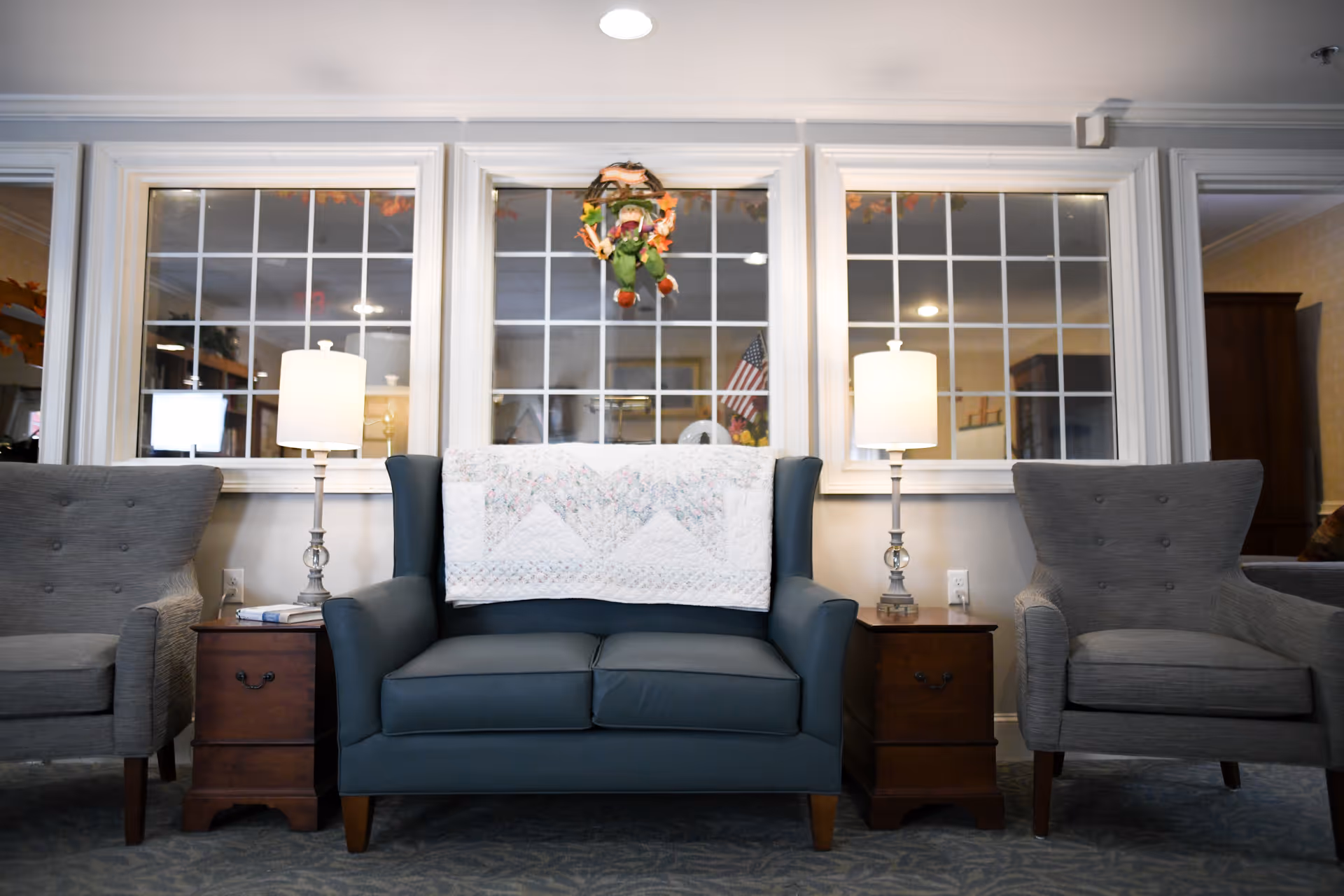 A seating area with a blue loveseat draped with a quilt, flanked by two gray armchairs and wooden side tables with lamps in front of multi-pane windows.