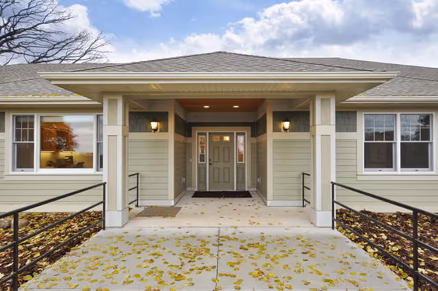 Front entrance of a single-story building with a covered porch, two wall-mounted lights, and a concrete walkway covered with fallen leaves. The building has light green siding with white trim and several windows.