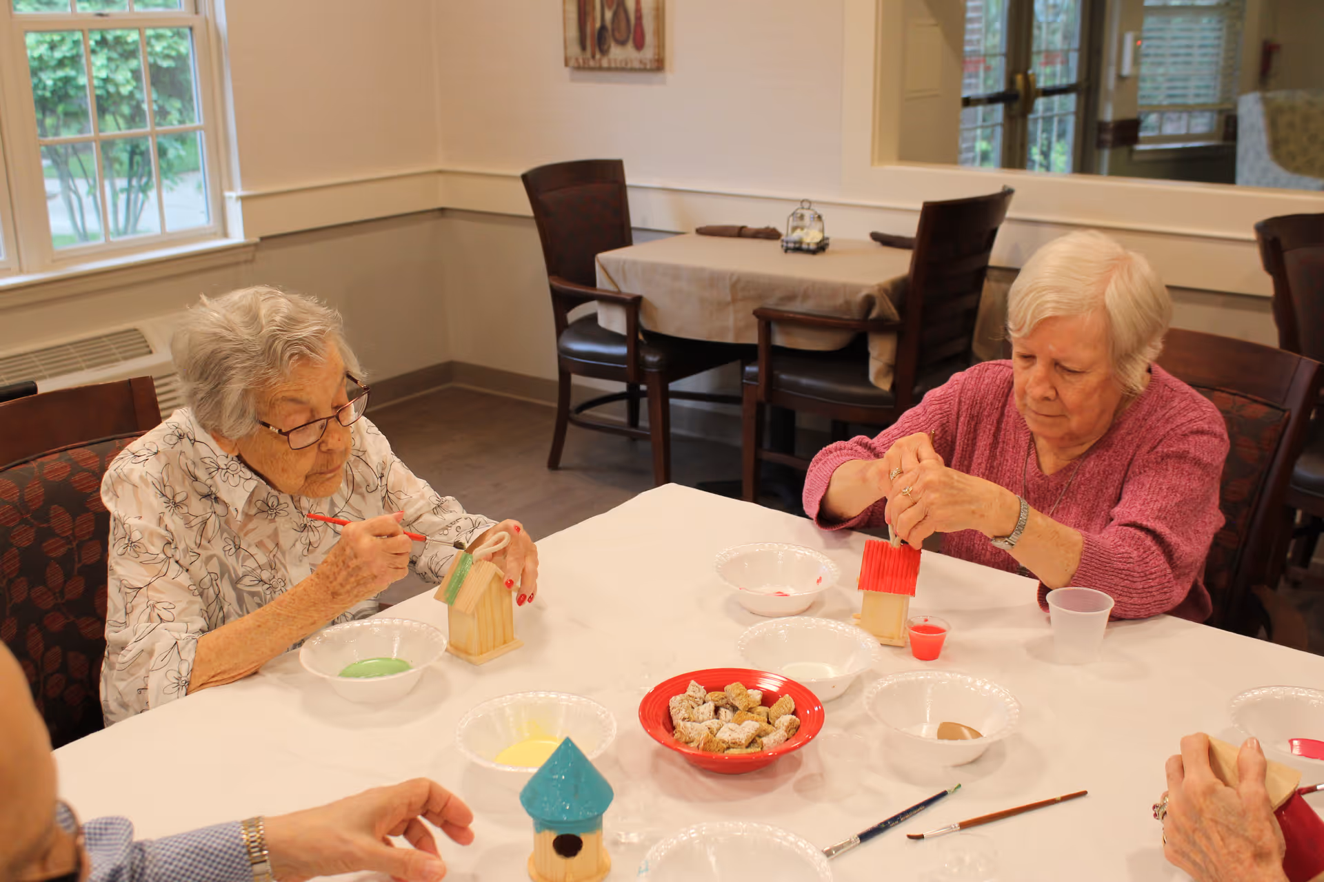 Two elderly women sitting at a table in a communal room, painting small wooden birdhouses with brushes. The table is covered with a white cloth and has several bowls of paint and a red bowl with snacks. The room has wooden chairs and a window with a view of greenery outside.