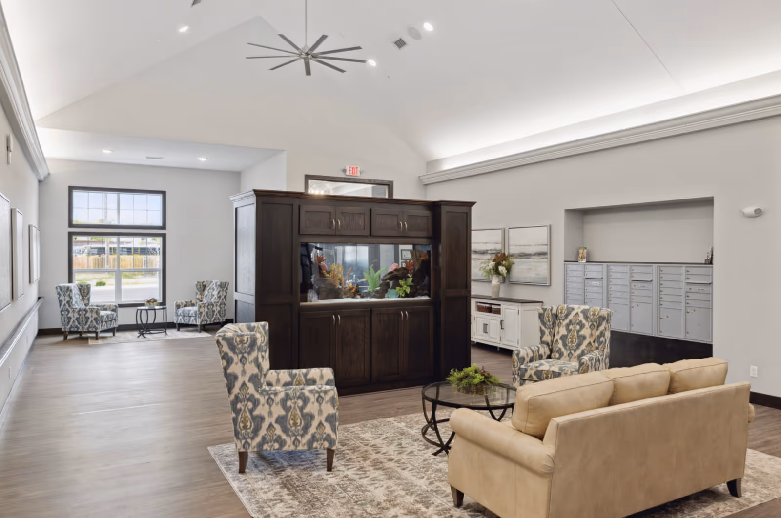 A spacious senior living facility common area with a large dark wood aquarium as a central divider. The room features a beige sofa, two patterned armchairs around a glass coffee table on a rug, and additional seating with two patterned chairs and a small table near a window. The walls are light gray with decorative paintings and a set of mailboxes recessed into one wall. The ceiling is vaulted with a large modern ceiling fan and recessed lighting.