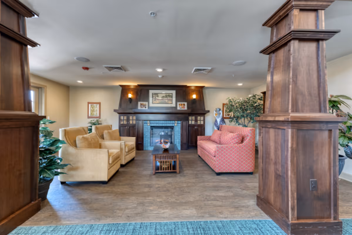 A cozy living room area with a wooden fireplace centered on the back wall, flanked by framed pictures and wall sconces. The room features two beige armchairs on the left and a red patterned sofa on the right, with a wooden coffee table in the middle. Large wooden columns frame the space, and there are several green plants around the room. The floor is wood, and the ceiling has recessed lighting.