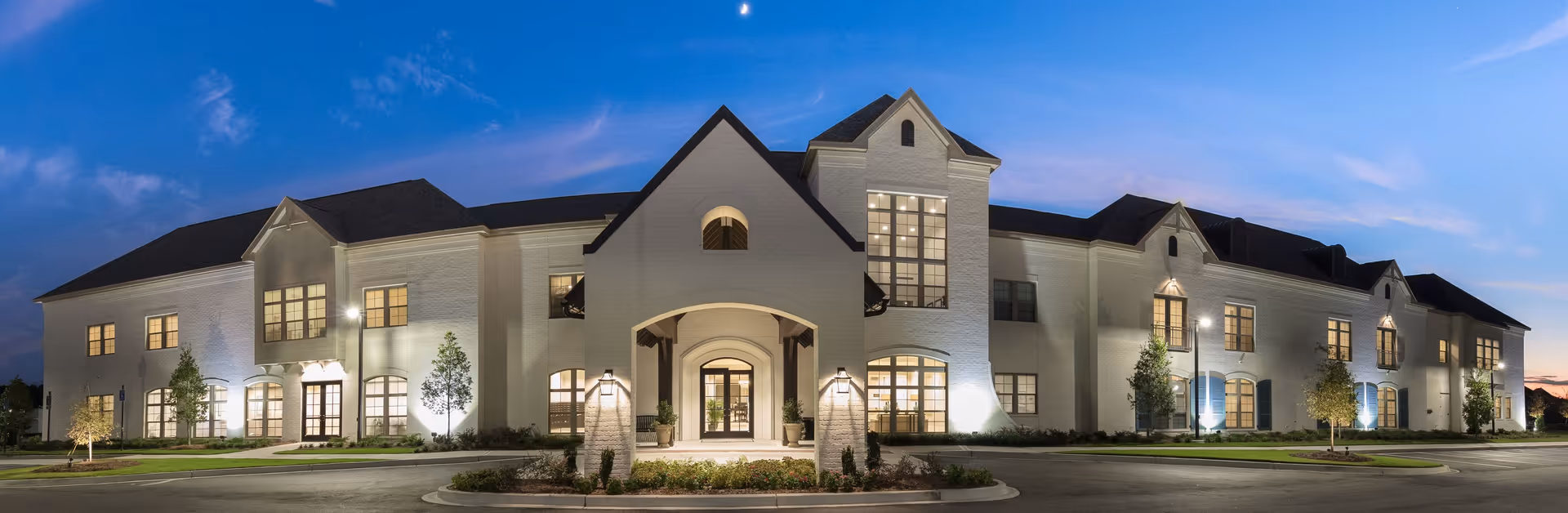 Panoramic view of a well-lit, two-story brick senior living facility with a central arched entrance, landscaped grounds, and a circular driveway at dusk.