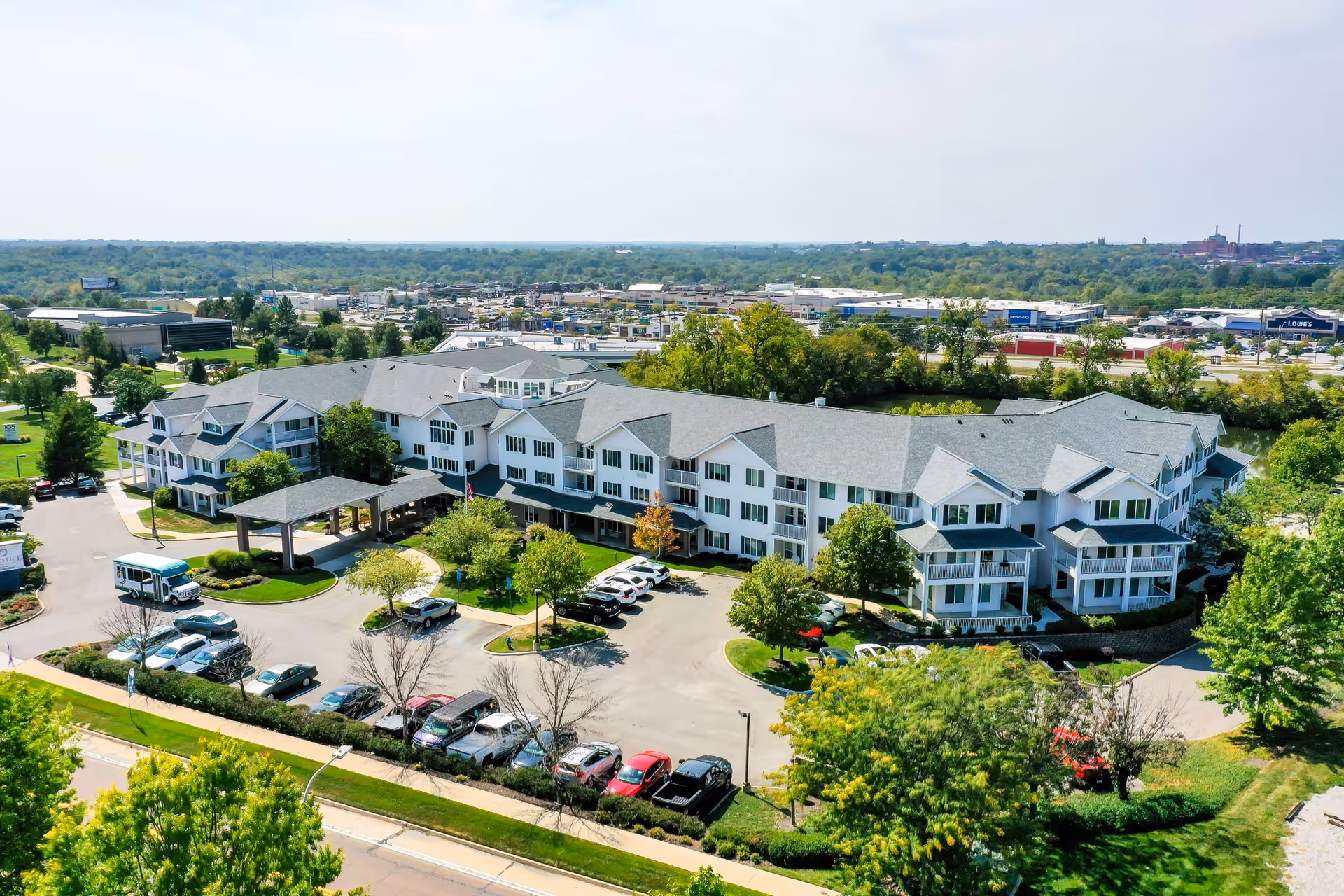 Aerial view of a large senior living facility building with multiple floors, surrounded by trees and parking lots with cars. The building has a gray roof and white exterior walls. The surrounding area includes greenery and a distant view of commercial buildings and a highway.