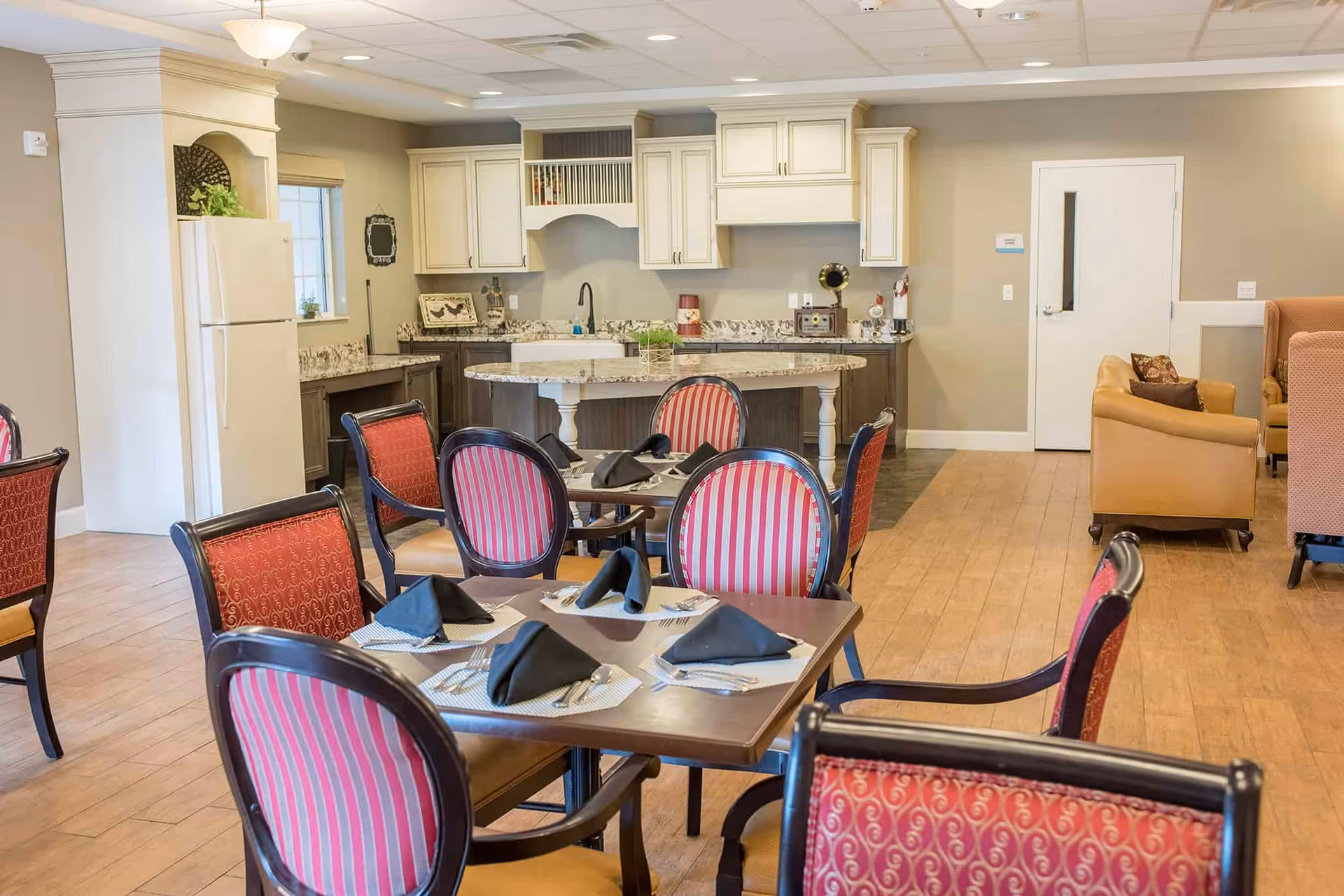 Communal dining area with set tables and red-striped chairs beside a kitchenette and lounge seating.