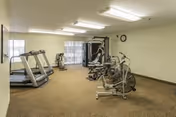 A fitness room with exercise equipment including two treadmills, a stationary bike, an elliptical machine, and a multi-function weight machine. The room has beige carpet, white walls, fluorescent ceiling lights, and a window with vertical blinds.