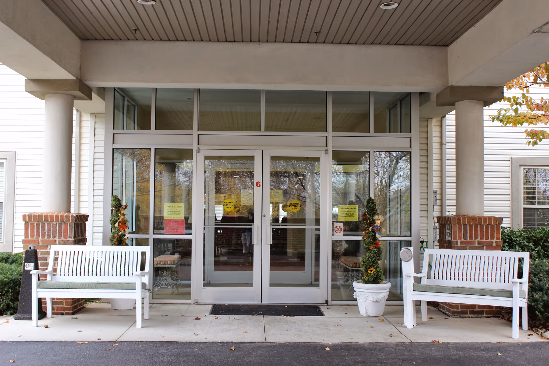 Glass double-door main entrance of a senior living facility with white benches and seasonal decorations.