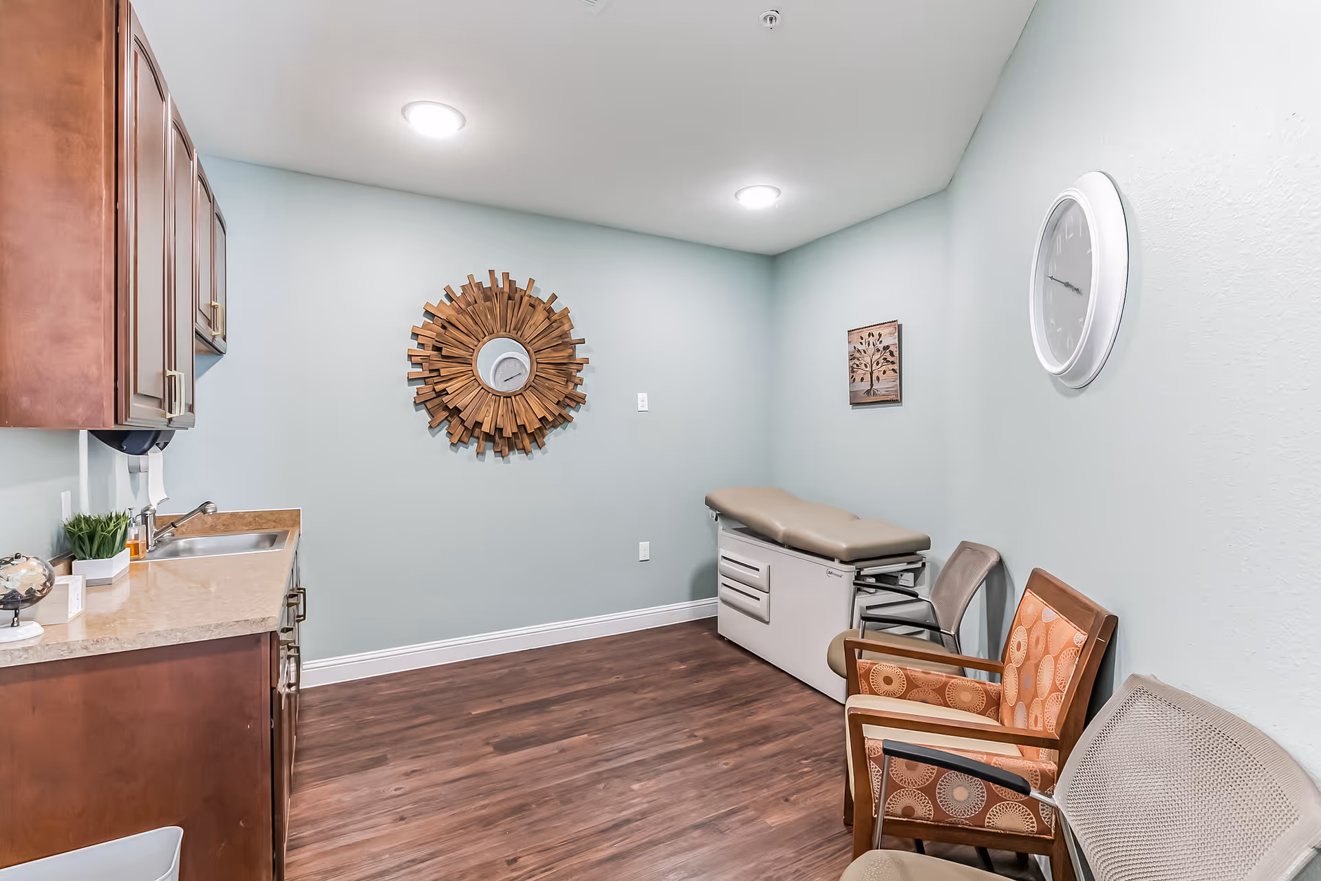 A small medical examination room with light blue walls and wood flooring. The room features a beige examination table against the far wall, three chairs with patterned upholstery along the right wall, a white clock on the right wall, a decorative wooden sunburst mirror on the back wall, and wooden cabinets with a countertop and sink on the left side.