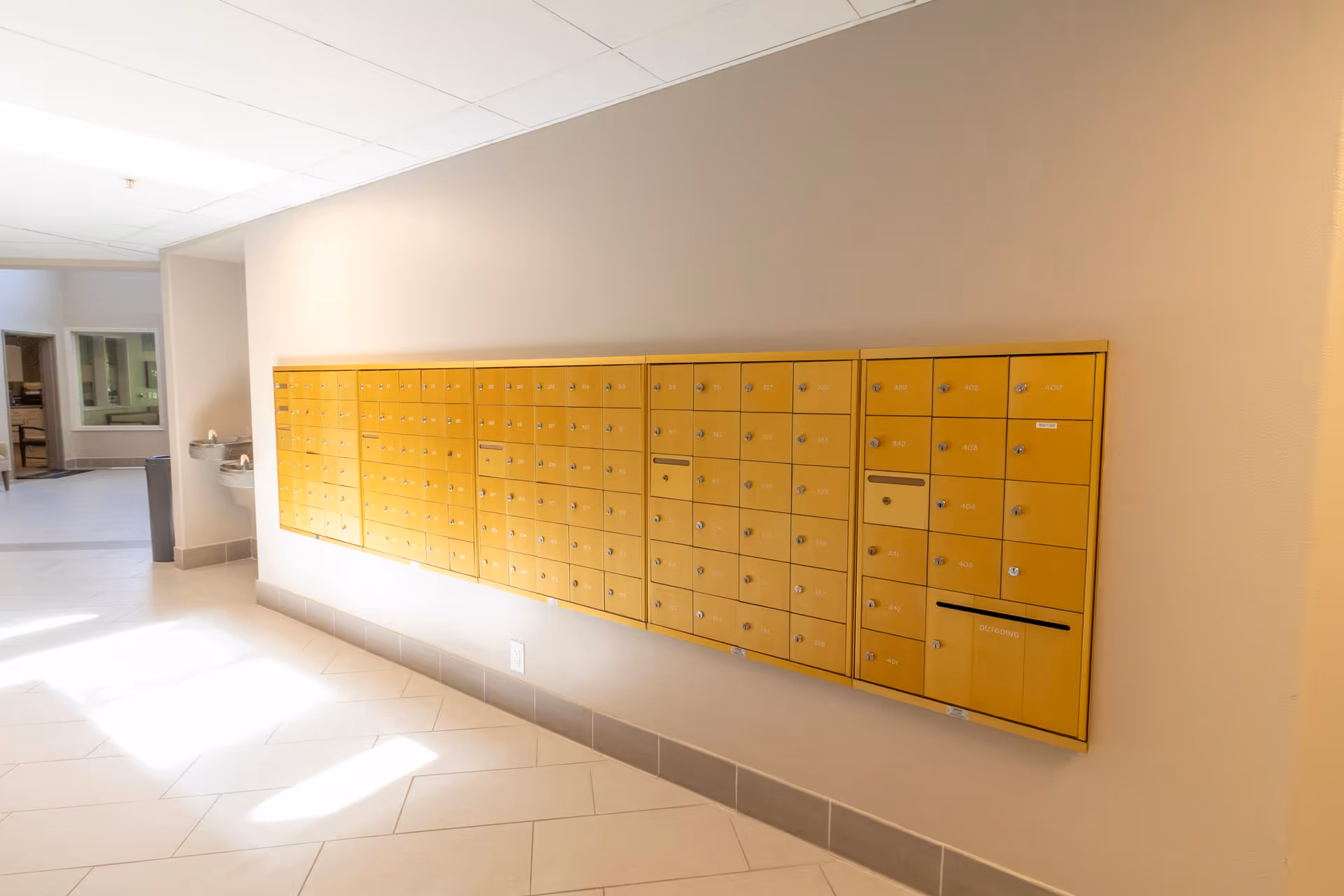 A hallway with a wall-mounted set of yellow mailboxes, a water fountain, and a trash can. The floor is tiled and the walls are painted beige.