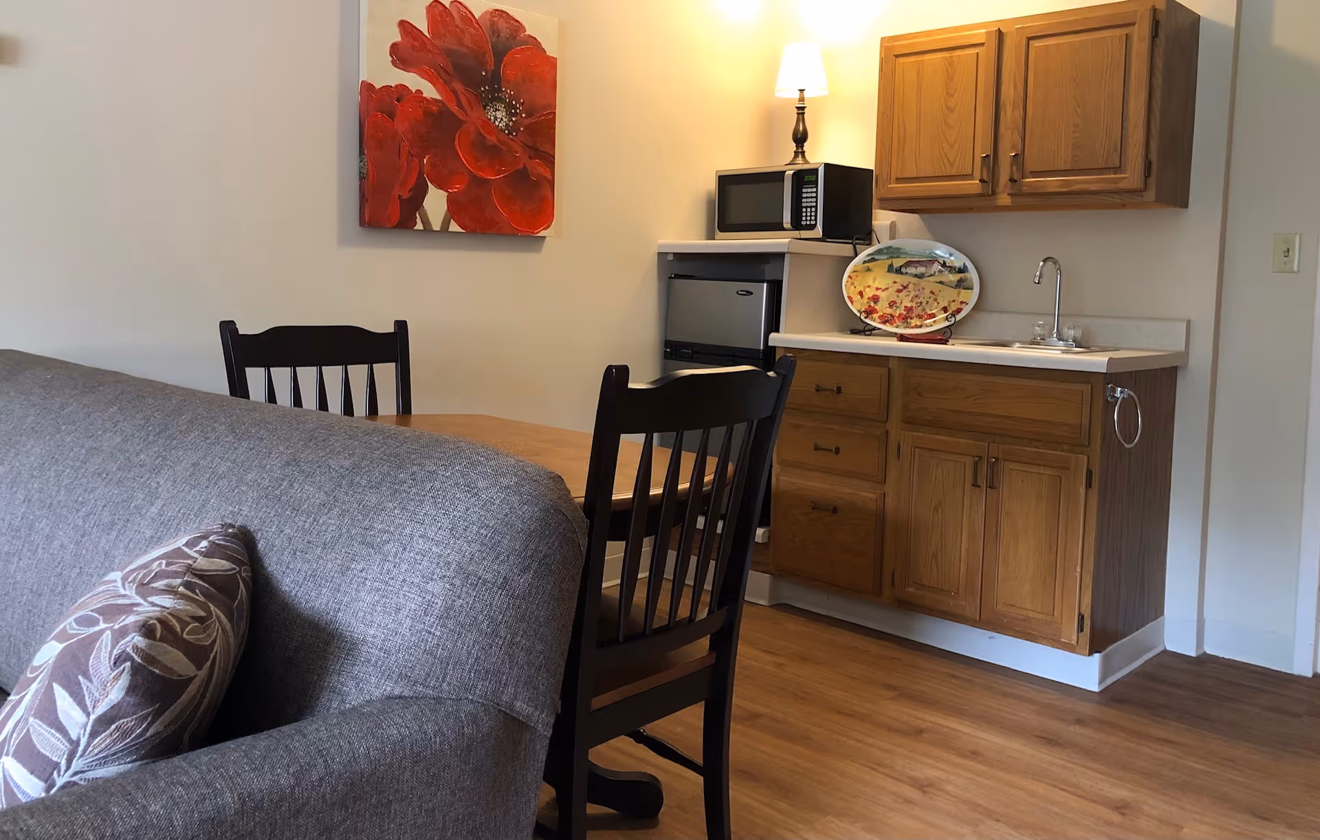 A small kitchenette area with wooden cabinets, a countertop with a sink, a microwave, and a mini refrigerator. In front of the kitchenette is a wooden dining table with two dark wooden chairs. A gray couch with a patterned pillow is partially visible in the foreground. A painting of red flowers hangs on the wall above the dining table.