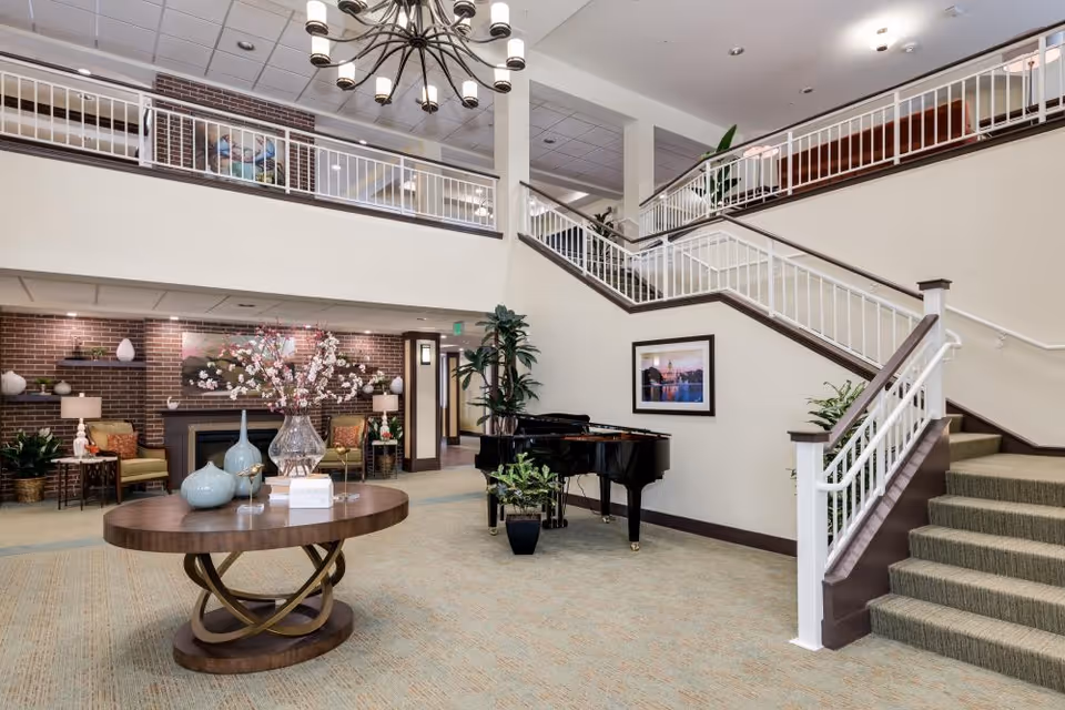 Spacious and well-lit interior common area with a grand piano, a round wooden table decorated with vases and flowers, a staircase with white railings leading to an upper floor, and a seating area with chairs and a fireplace against a brick wall.