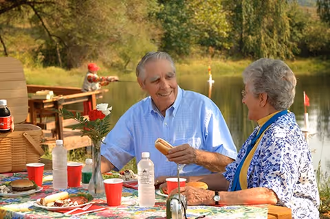 An elderly man and woman sitting at a picnic table outdoors near a lake, enjoying a meal together with plates of food, drinks, and a vase with flowers on the table. The man is holding a hot dog and smiling at the woman, who is also smiling back. Trees and water are visible in the background.