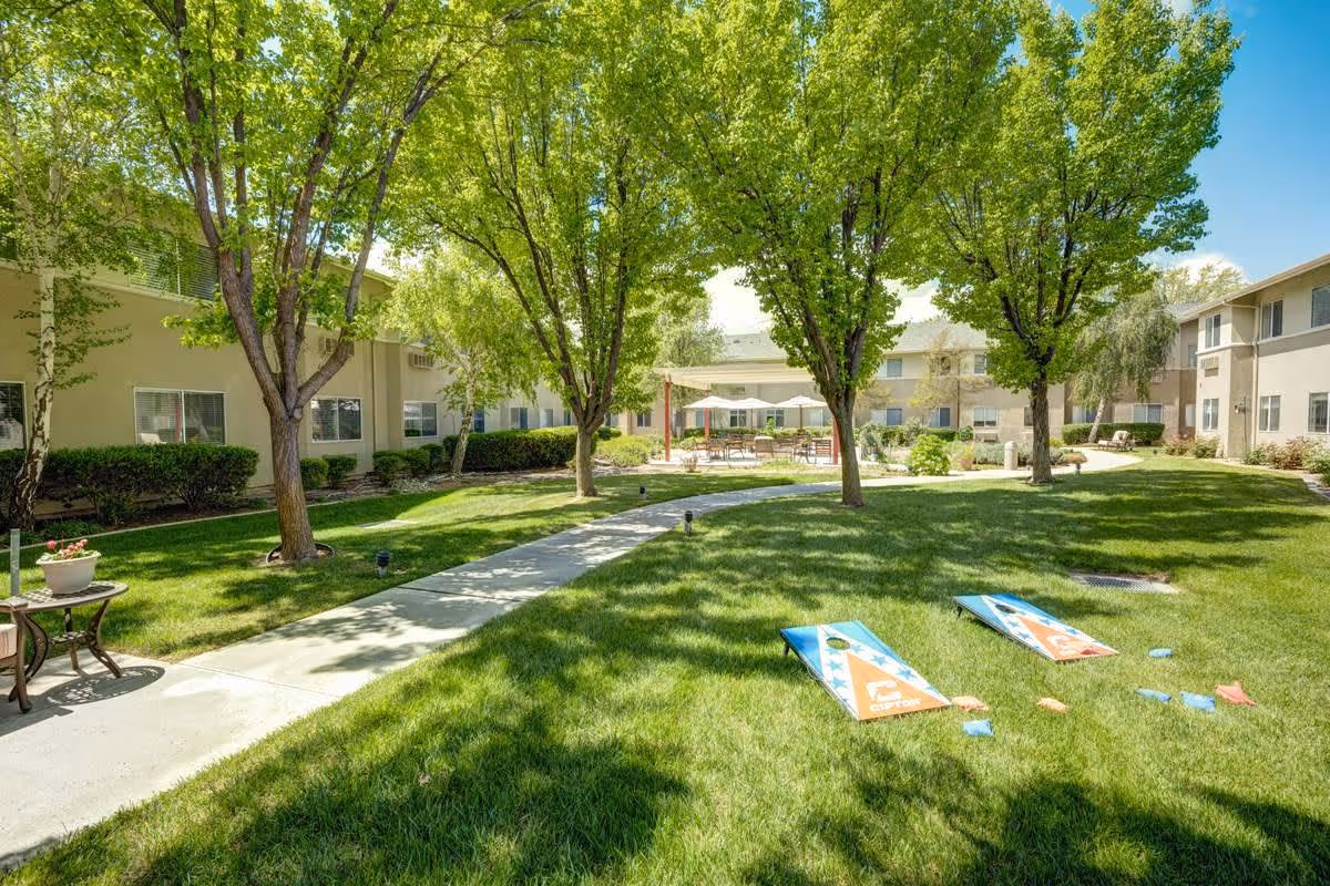 Outdoor courtyard area at Creston Village with green grass, several trees, a concrete walkway, and two cornhole boards set up on the lawn. Surrounding the courtyard are two-story residential buildings with windows. There are tables and chairs under umbrellas in the background.