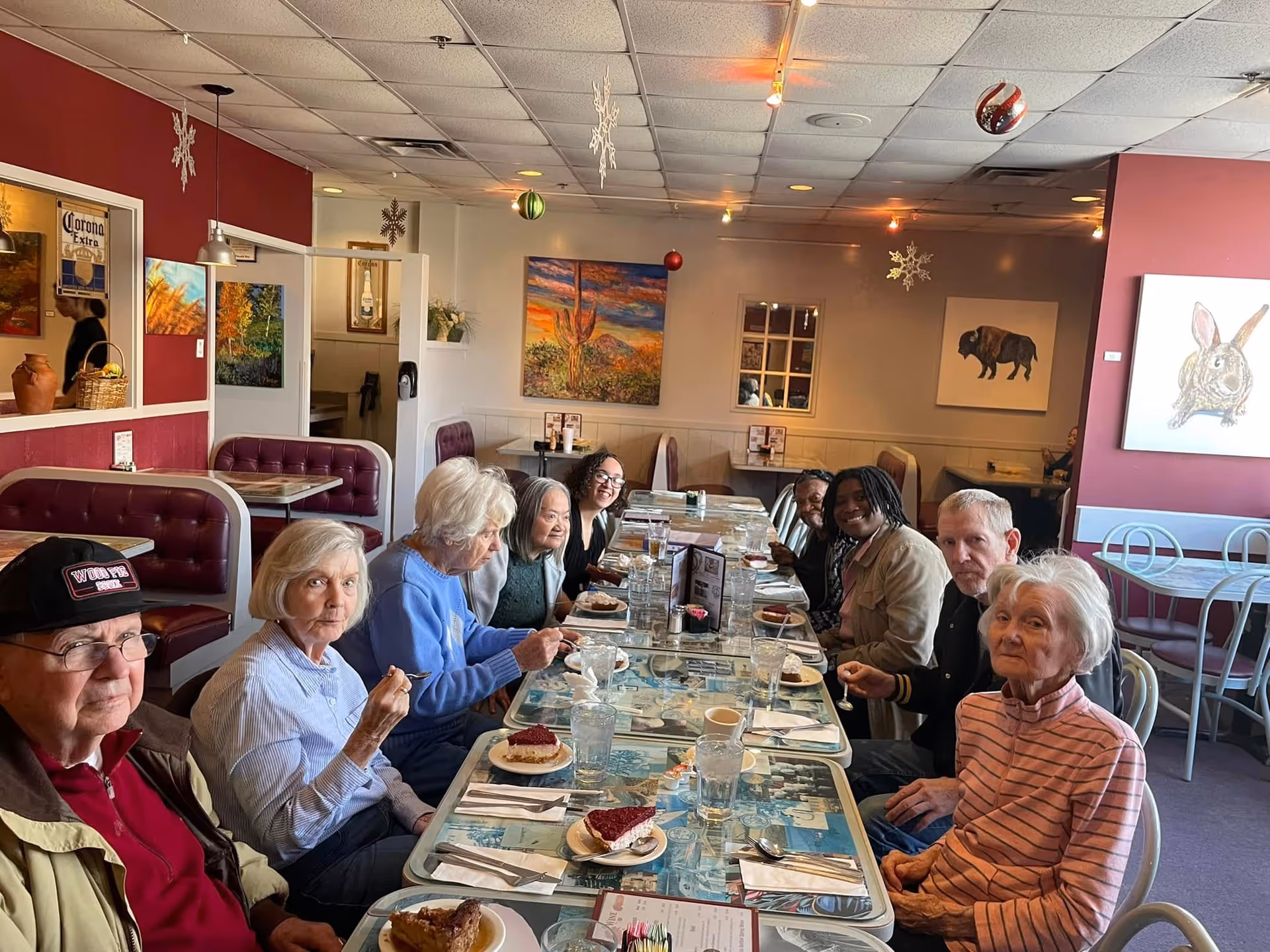 A group of elderly people and two younger adults sitting around a long table in a restaurant, enjoying slices of pie and drinks. The restaurant has red walls decorated with paintings of a cactus, a bison, and a rabbit, as well as hanging holiday ornaments and snowflakes. The seating includes red cushioned booths and metal chairs.