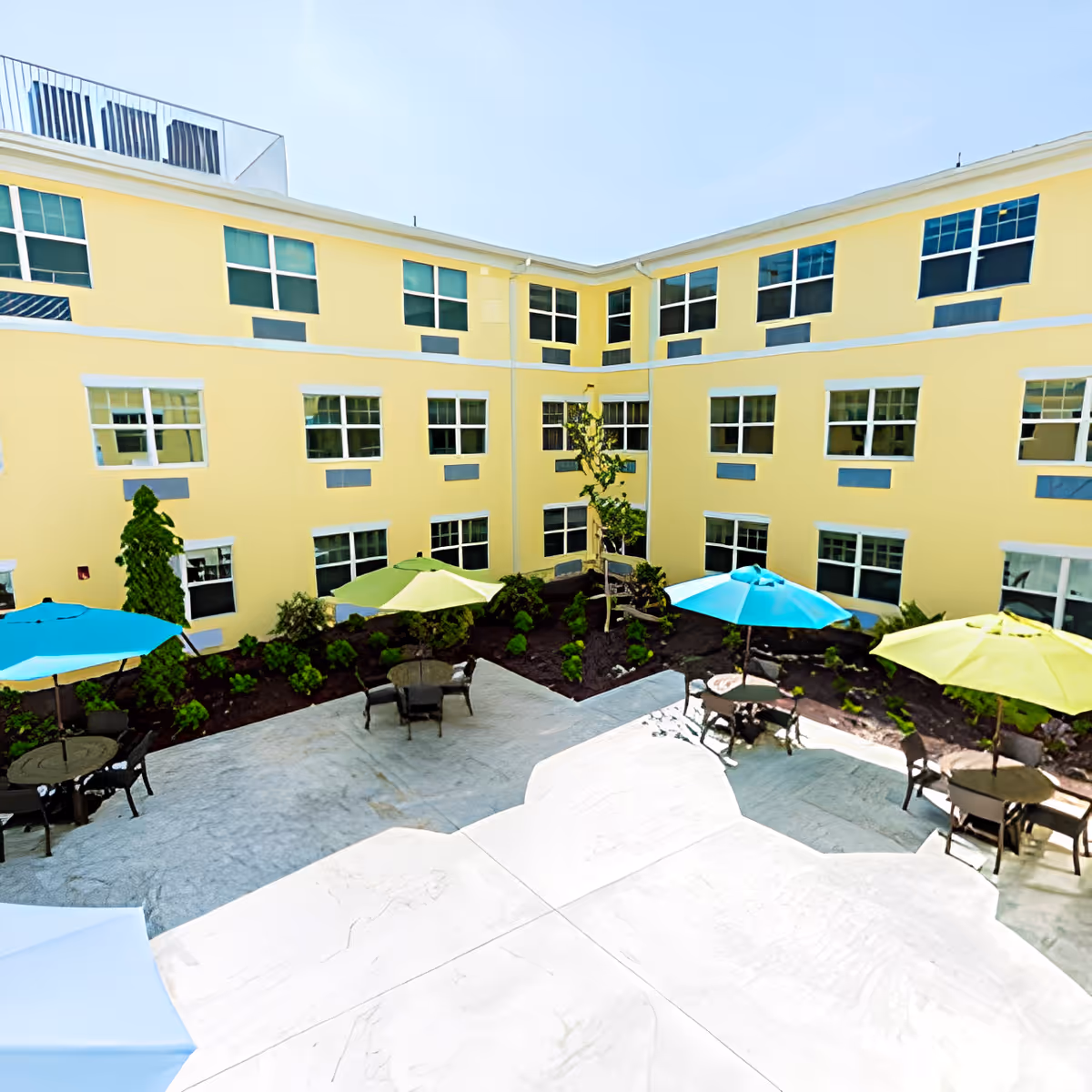 Outdoor courtyard area of a senior living community with yellow building walls, multiple windows, and several patio tables with colorful umbrellas in blue, green, and yellow. The courtyard has landscaped garden beds with small bushes and a young tree.