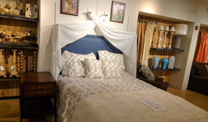 A furnished bedroom display featuring a patterned bed with pillows, a nightstand, and shelving with lamps and vases in an interior space.