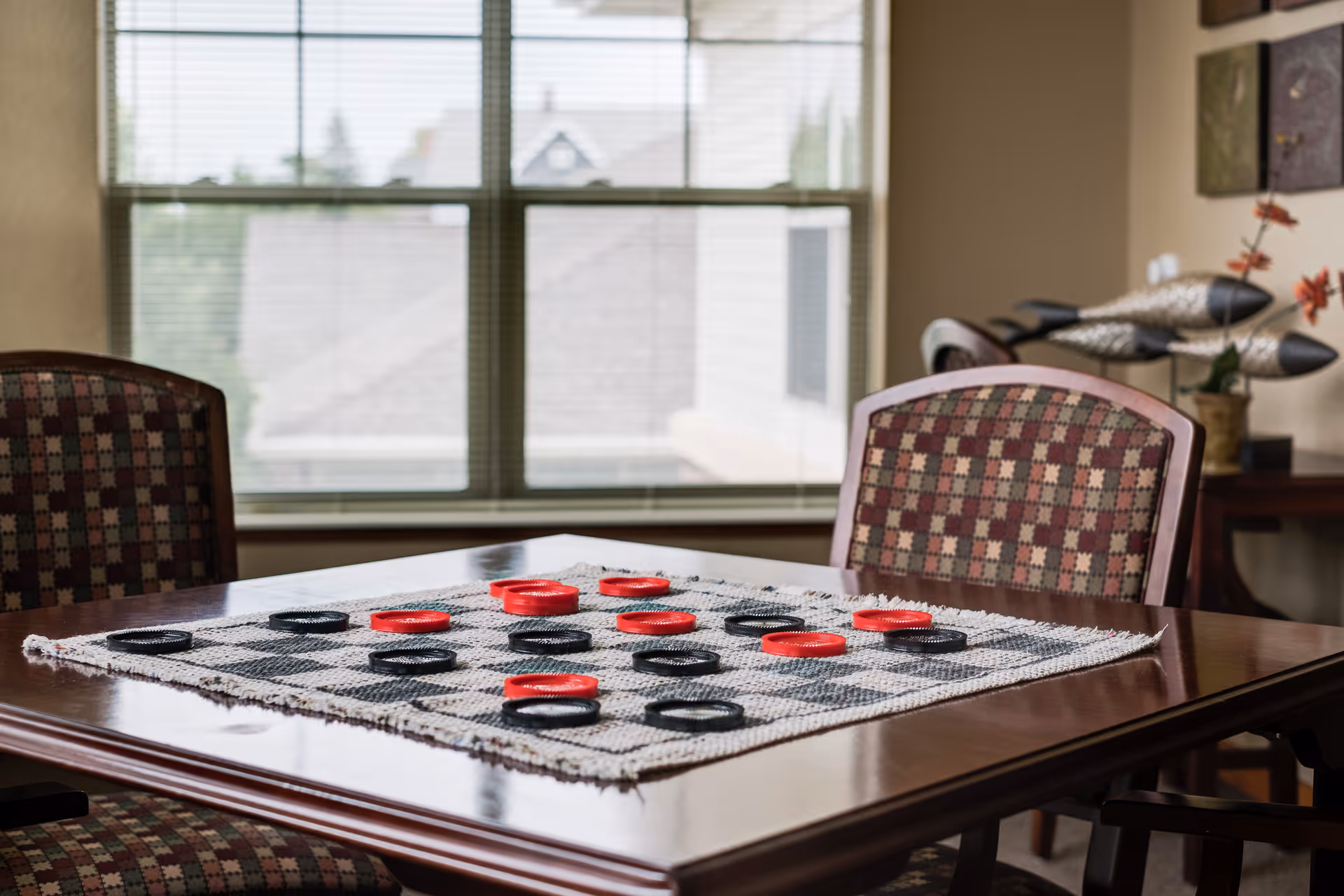A wooden table with a checkers game set up on a woven checkerboard cloth. The table is surrounded by patterned cushioned chairs. In the background, there is a large window with blinds and some decorative items on a side table.