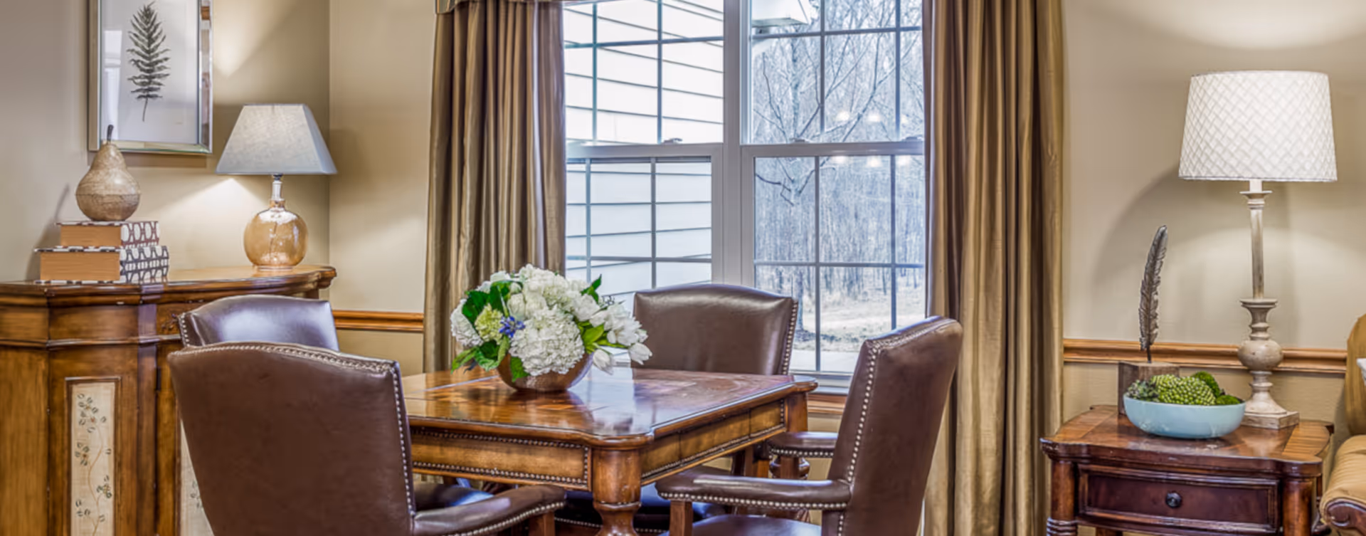 A cozy dining area with a wooden table surrounded by four brown leather chairs. On the table is a floral centerpiece with white and green flowers. Behind the table is a large window with beige curtains, letting in natural light. To the left is a wooden cabinet with a lamp and decorative items, and to the right is a wooden side table with a lamp and a decorative bowl.