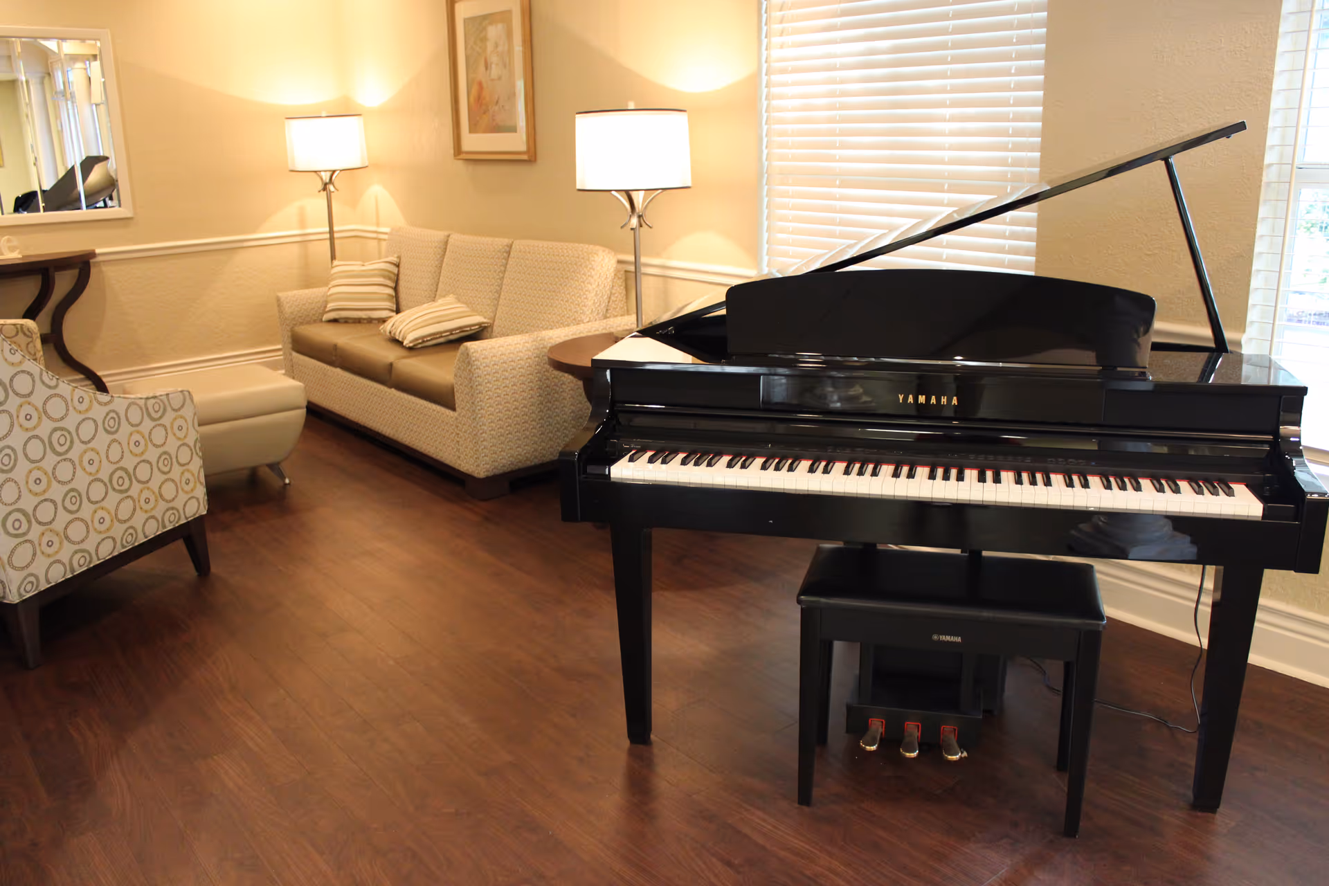 Bright lounge area with a black Yamaha grand piano in the foreground and a sofa, armchair, and lamps in the background.