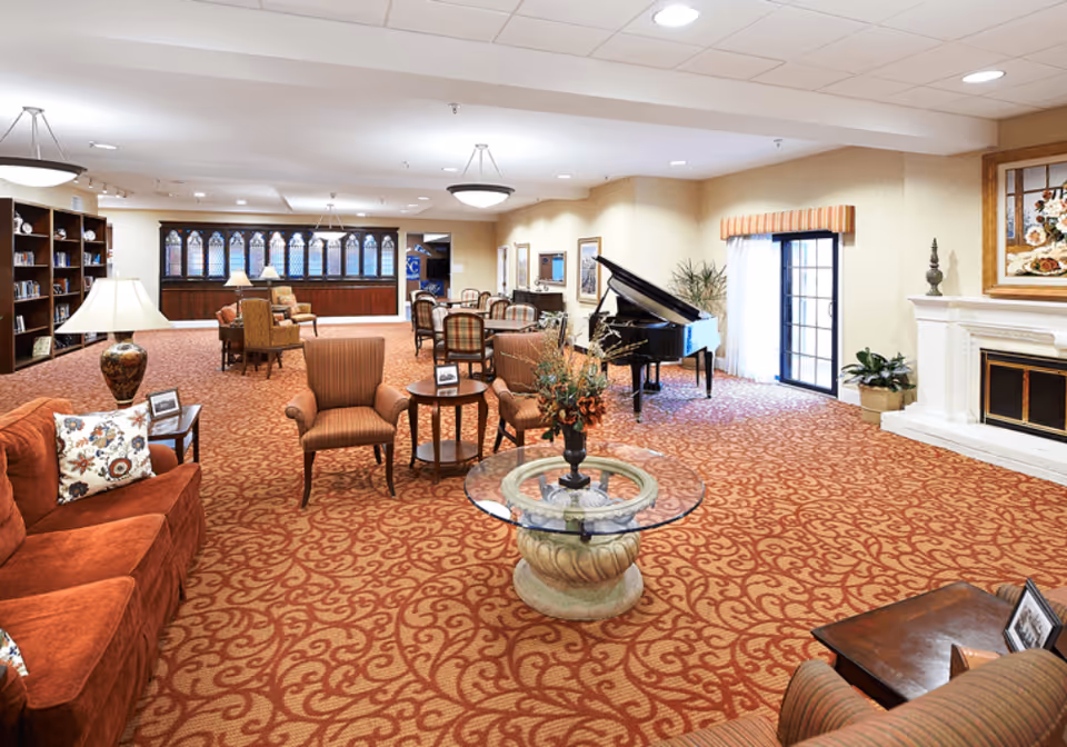 A spacious senior living facility common area with patterned carpet, multiple seating arrangements including armchairs and sofas, a glass-top round table with a floral centerpiece, a grand piano near a sliding glass door, a fireplace with a floral painting above it, and bookshelves along the back wall.