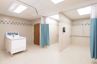 A clean, tiled room with multiple shower stalls separated by partial walls and blue privacy curtains. There is a white washing machine on the left side and a wooden door in the background. The walls have a decorative green and white tile border near the ceiling.